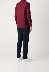 Bordowa koszula z długim rękawem z detalami guzików, w połączeniu z granatowymi spodniami i białymi trampkami. Gładka tkanina, dopasowany krój i casualowy styl.