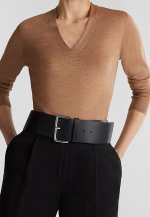 Person iført en kamelfarvet V-hals sweater, stoppet ned i sorte bukser med et bredt sort bælte og sølvsæt spænde, hænderne i lommerne.