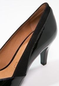 Chaussure à talon haut en cuir verni noir avec garniture en daim et intérieur en cuir beige, présentant un bout rond et un talon de hauteur moyenne.