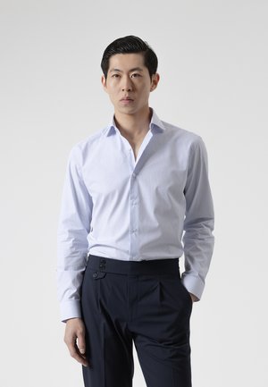 Joven con el pelo peinado hacia atrás que lleva una camisa de vestir azul claro con rayas metida en pantalones oscuros a medida.