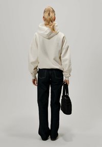 Sudadera blanca oversized con capucha, combinada con pantalones vaqueros oscuros de pernera acampanada. El modelo sostiene un pequeño bolso negro con una correa.