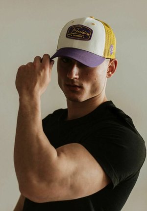 White and purple cap with a yellow mesh back, featuring a rectangular logo patch. The person is holding the cap, showing muscular arm details.