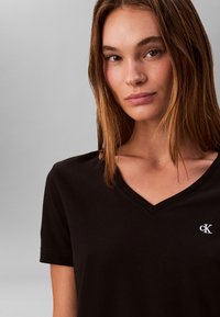 T-shirt in cotone nero con scollo a V, maniche corte e piccolo logo bianco "cK" sul lato sinistro. Tessuto morbido e design minimalista.