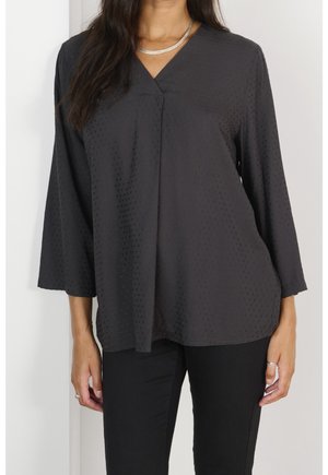 Femme portant un chemisier noir à col en V avec une texture subtilement ponctuée et des manches trois-quarts, associé à un pantalon noir et un collier en argent.