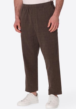 Uomo con pantaloni marroni a vita elasticizzata e texture e sneakers bianche, in piedi davanti a uno sfondo semplice.