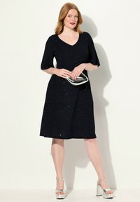 Robe noire avec des manches courtes, col en V et texture scintillante. Associée à des talons compensés argentés et une pochette métallique.