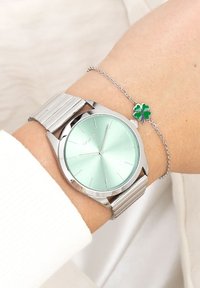 Montre argentée avec un cadran vert menthe et un bracelet en métal argenté, associée à un mince bracelet argenté avec un charm en trèfle à quatre feuilles vert.