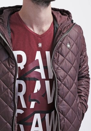 Homme portant une veste matelassée bordeaux sur un T-shirt graphique assorti bordeaux avec un texte blanc et noir, debout avec une main dans la poche.