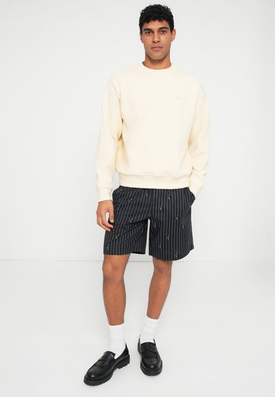Sweatshirt crème, shorts noirs rayés avec de petits motifs, chaussettes blanches et chaussures noires. La tenue présente une coupe décontractée et un style informel.