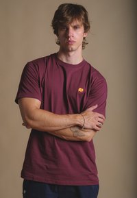 T-shirt di cotone color bordeaux con maniche corte, presenta un piccolo logo di un elefante arancione sul lato sinistro del petto. Texture liscia e vestibilità standard.