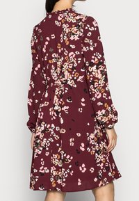 Burgundy klänning med lång ärm, prydd med ett blommigt mönster i rosa, vitt och svart. Samlad midjedetalj och flödande kjol.