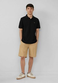 Schwarzes Polohemd mit kurzen Ärmeln, kombiniert mit beigen Shorts und beigen Sneakern. Einfaches Design mit minimalen Akzenten.