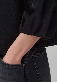 Schwarze Bluse mit Puffärmel, aus einem glatten Material gefertigt. Eine Hand liegt in einer Tasche von dunkelgrauen Denim-Jeans mit einem kleinen metallischen Knopf.