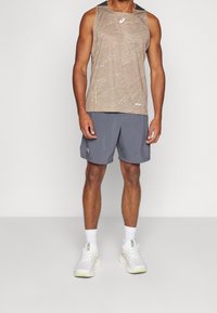 Uomo che indossa un top senza maniche beige con motivo, pantaloni corti grigi da ginnastica, calze bianche e scarpe da corsa bianche, in piedi contro uno sfondo semplice.