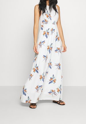 Nainen pukeutuneena valkoiseen, hihattomaan maksi-mekkoon, jossa on sininen ja oranssi kukkakuvio, sekä mustiin sandaaleihin, seisoo yksinkertaisen taustan edessä.