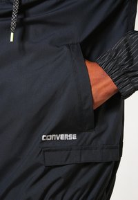 Svart vindjacka med elastiska ärmmuddar, med en framficka och en silverfärgad "Converse"-logotyp. Tyget är slätt med en lätt lyster.