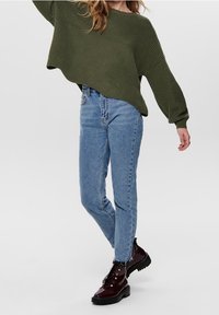 Pull côtelé vert olive, jean taille haute bleu clair, et bottines en cuir verni bordeaux avec semelles noires.