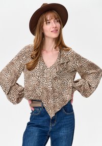 Kobieta z długimi brązowymi włosami, ubrana w brązowy kapelusz, bluzkę w panterkę z falbankami oraz niebieskie dżinsy, stojąca z rękami na biodrach.