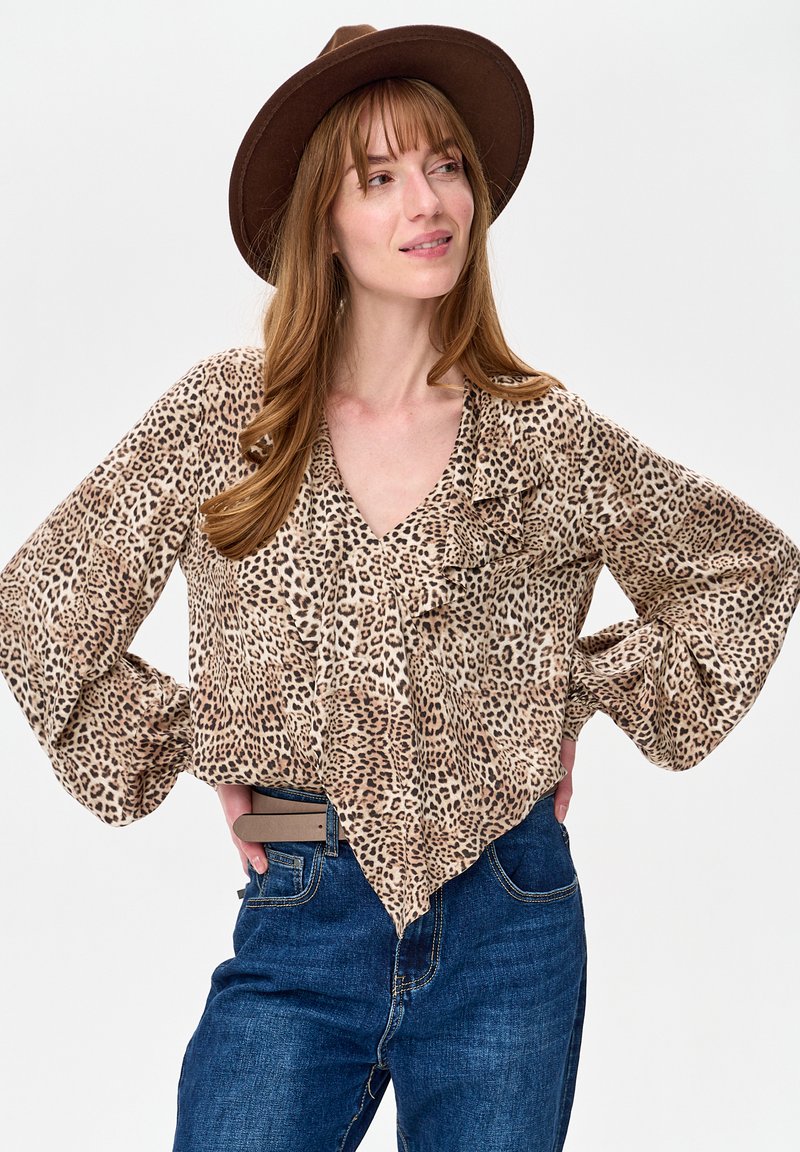 Kobieta z długimi brązowymi włosami, ubrana w brązowy kapelusz, bluzkę w panterkę z falbankami oraz niebieskie dżinsy, stojąca z rękami na biodrach.