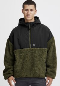 Olivgrön fleece-tröja med svart nylonöverkropp. Har en huva, halv dragkedja och en liten varumärkespatch på bröstet. Strukturerad tyg.