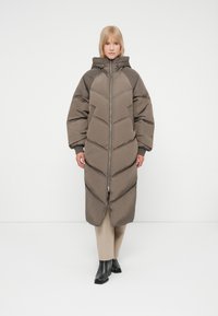 Abrigo de plumas en color topo con patrón de rombos, capucha oversized y cremallera delantera. La modelo lleva pantalones beige y botas negras.