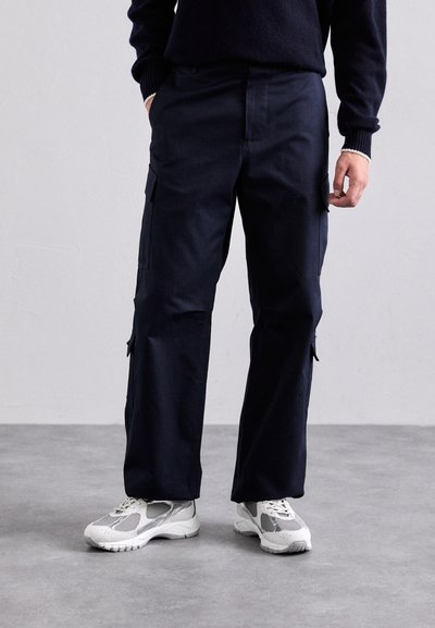 Pantaloni cargo navy cu o croială lejeră, dotati cu buzunare laterale, o textură fină a stofei și picioare ușor conice, asortați cu adidași albi.