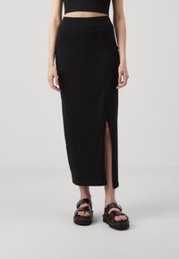 Svart, knälång kjol med en sidoklyfta, tillverkad av ett slätt tyg; kombineras med kraftiga, mörka sandaler som har flätade remmar och en tjock sula.