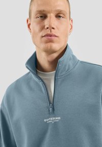 Felpa azzurro chiaro con zip e collo alto, con un logo bianco sul petto. Tessuto morbido e elementi di design moderni.
