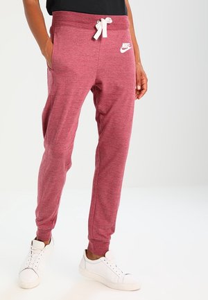 Personne portant un pantalon de survêtement Nike bordeaux avec un cordon blanc et des baskets blanches, debout les mains dans les poches.