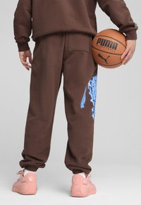 Hnedé tepláky s manžetami na členkoch majú modrý grafický dizajn. Model drží hnedý basketbal a má na sebe ružové tenisky.