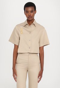 Beige, rövid ujjú cropped ing ezüst gombokkal és sárga hímzéssel a bal mellrészen. Az anyag simának és kissé textúráltak tűnik.