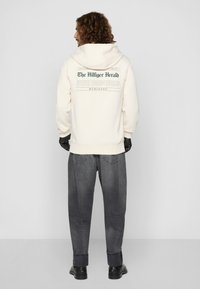 Kremkrāsas džemperis ar kapuci, uz kura aizmugures ir zaļa teksta druka. Apvienots ar brīviem pelēkiem džinsiem un melniem apaviem.