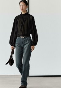 Sort gennemsigtig bluse med rynket halsudskæring, brede ærmer; blå jeans med lige ben; brun taske; sølvfarvet bæltespænde; minimalistisk miljø.