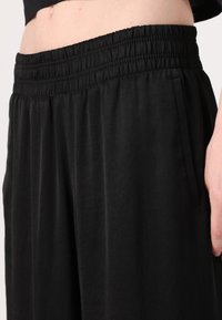 Black, loose-fitting pants with an elastic waistband, featuring side pockets and a smooth, lightweight texture. No visible patterns.