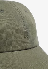 Gorra verde oliva con tela texturizada, que presenta un sutil diseño bordado de calavera y huesos cruzados en el frente. Forma clásica de visera curvada.