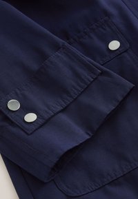 Marineblaues Stoffbündchen mit zwei silbernen Druckknöpfen auf einer abgesteppten Taschenklappe eines Kleidungsstücks.