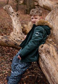 Green padded jacket with faux fur trim, silver accents, and a logo on the sleeve. Gray patterned pants with a loose fit. Engaged in nature.