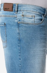 Jeans de mezclilla azul claro con una textura suave, diseño de cinco bolsillos, parche de cuero marrón en la cintura y costura en contraste.