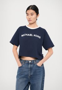 T-paita laivastonsinisestä kankaasta, jossa on valkoinen pyöreä kaula-aukko ja "MICHAEL KORS" -logo lihavilla kirjaimilla edessä. Kangas vaikuttaa sileältä ja kevyeltä.