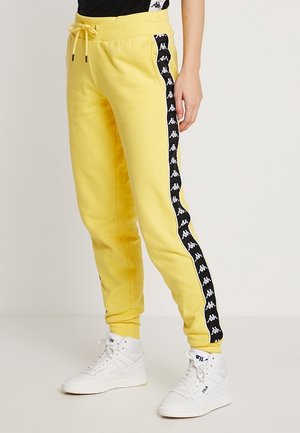 Gule joggingbukser med sort-hvide logo-striber ned langs siderne, båret med hvide high-top sneakers og en sort top.