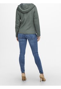 Grön huva med fleecefoder, som har en figurnära passform, i kombination med slim-fit blå denimjeans och beige högklackade skor.