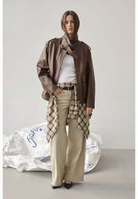 Frau trägt übergroße braune Lederjacke, weißes Oberteil, beige Hosen mit weitem Bein, karierte Hemd um die Taille gebunden und schwarze spitze Schuhe, steht drinnen.