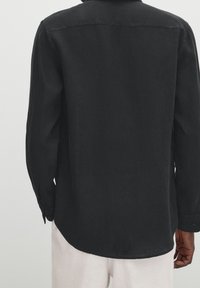 Zwart lange mouwen shirt van een gestructureerde stof, met een gebogen zoom en knopenmanchetten, weergegeven vanaf de achterkant. Eenvoudig ontwerp.