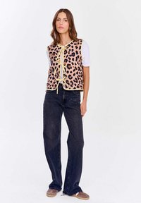 Žena v béžovo-černém vestě s leopardím potiskem přes bílou košili, tmavých širokých kalhotách a hnědých teniskách, stojící na bílém pozadí.