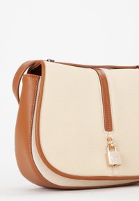 Bolsa de lona beige con acentos de cuero marrón, de forma redondeada, tapa superior y un pequeño detalle de candado. Textura suave y correa ajustable.