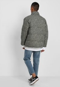 Kurtka puchowa w szarym kolorze z czarnymi cętkami w panterkę. Posiada wysoki kołnierz i pikowany wzór. Nosi się ją z białą koszulą i luźnymi niebieskimi dżinsami.