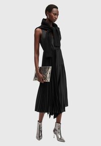 Mustat laskostetut mekko ilman hihoja, yhdistetty mustaan neuleeseen, joka on kiedottu olkapäiden ympärille, ja hopeisiin nilkkureihin; pitämässä metallista clutch-laukkua.