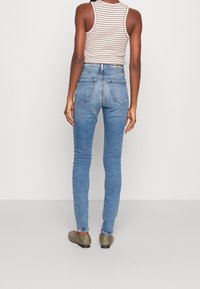 Pantalones vaqueros ajustados de cintura alta y dos bolsillos traseros, combinados con una camiseta de tirantes a rayas y zapatos planos de color oliva.
