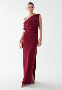 Burgundijas, vienas pleca maxi kleita ar plūstošu audumu, pieguļošu siluetu un sānu izgriezumu; nēsāta ar zelta aksesuāriem un siksnām sandalēm.