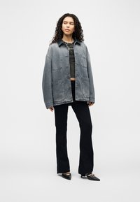 Femme aux cheveux bouclés portant une veste en jean délavée oversize, un haut court noir, un pantalon noir et des chaussures noires pointues, debout sur un fond blanc.